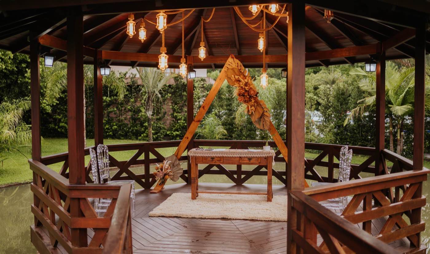 Interior do gazebo de madeira com lanternas pendentes e arco rústico de flores secas, cercado pelo jardim