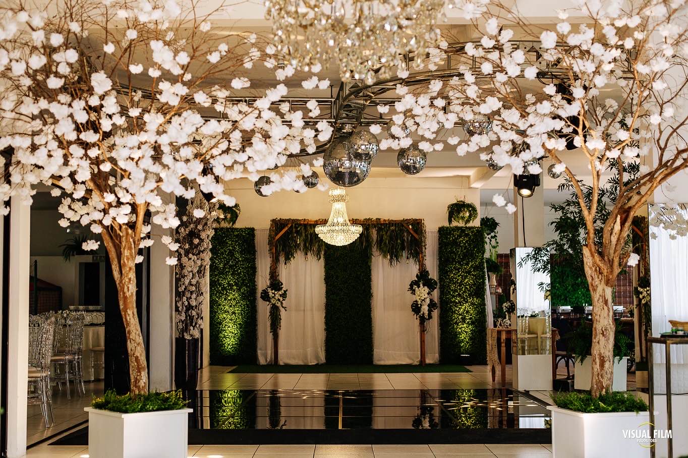 Salão climatizado decorado com árvores de flores brancas, lustre de cristal e pista de dança espelhada