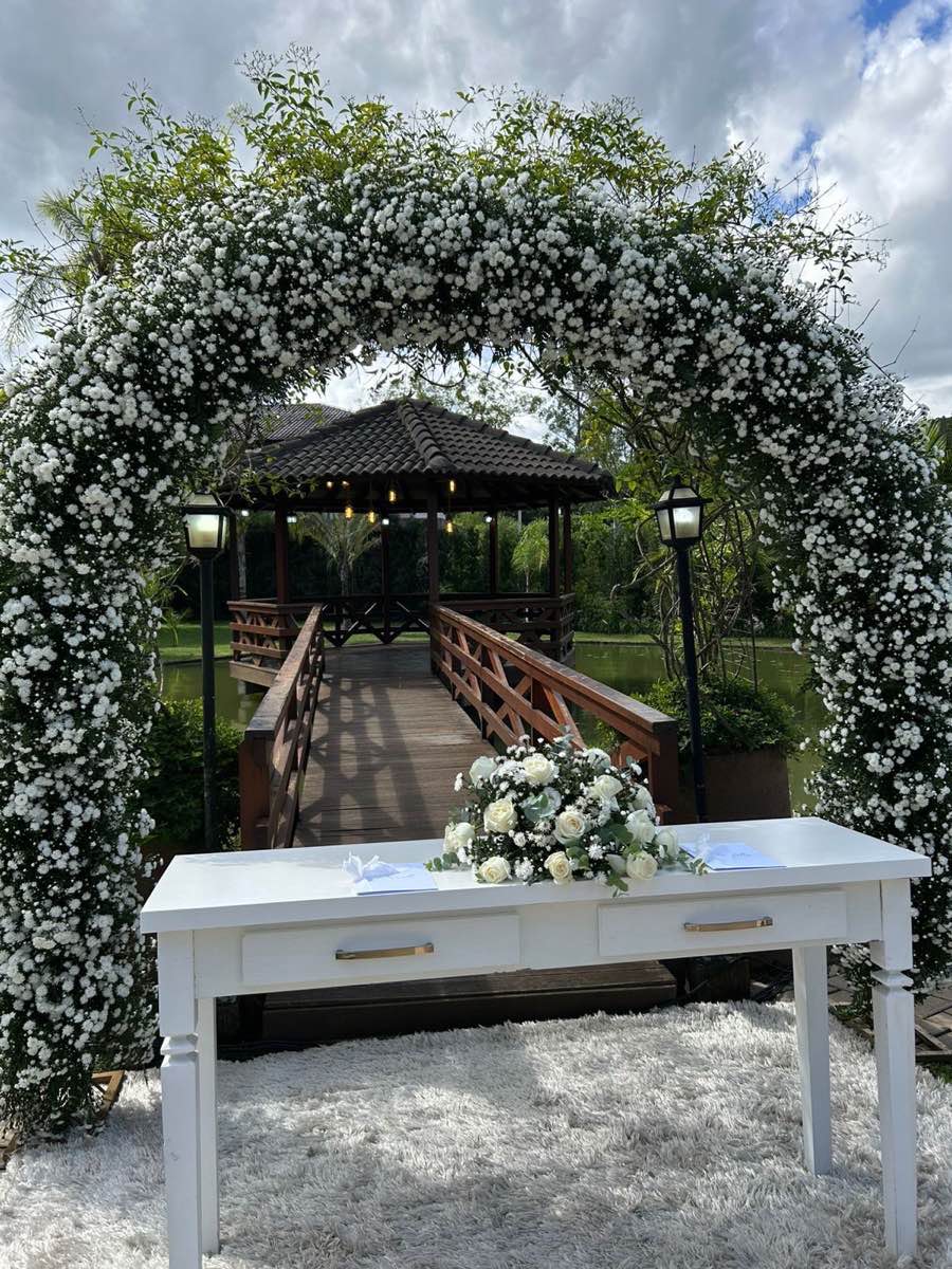 Arco de flores brancas sobre a mesa do celebrante, com a ponte de madeira que leva ao gazebo sobre o lago