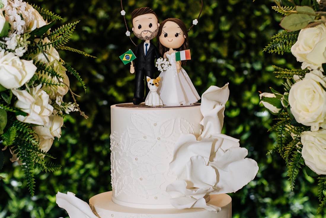 Bolo de casamento branco com topo de noivinhos, entre arranjos de rosas brancas e folhagens