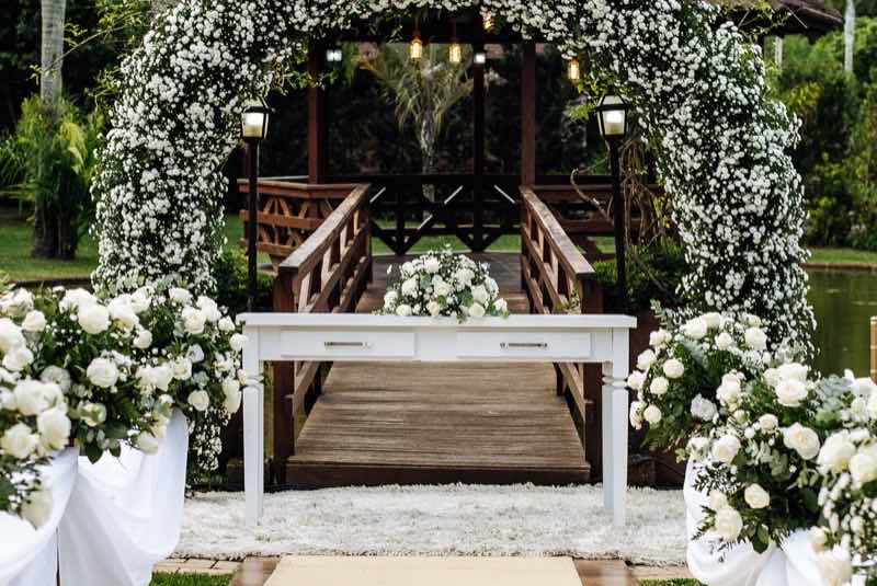 Mesa do celebrante sob arco de flores brancas iluminado por lanternas, diante da ponte do gazebo
