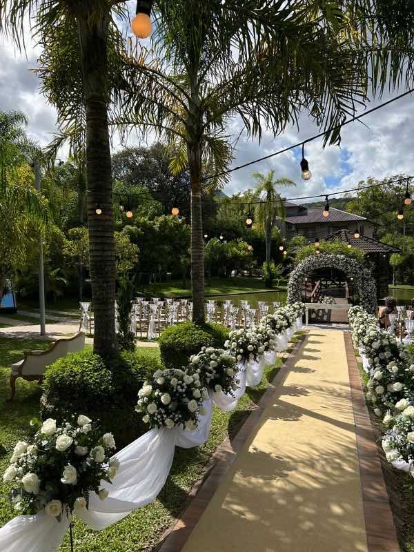 Corredor da cerimônia sob palmeiras em dia de sol, com as cadeiras dos convidados e o arco de flores ao fundo
