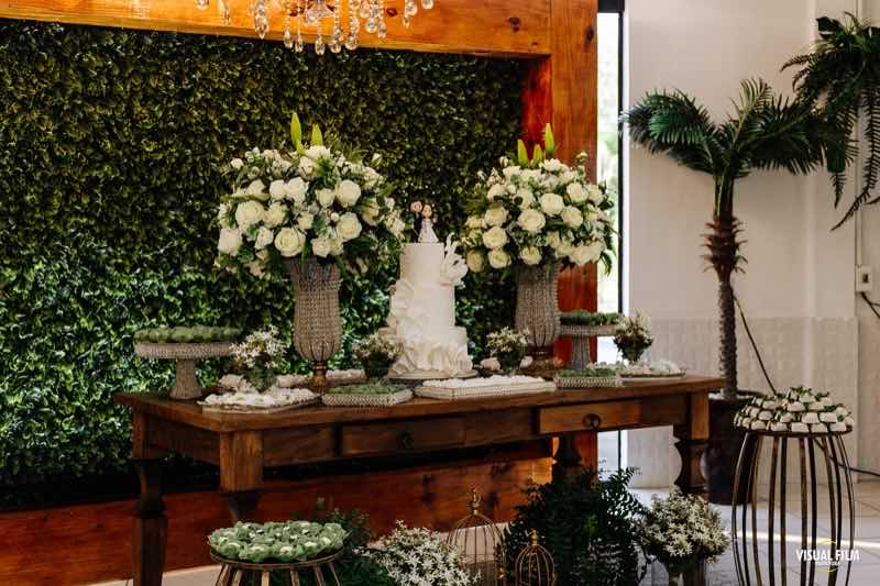 Mesa do bolo em madeira rústica com arranjos de rosas brancas diante de um painel de folhagens