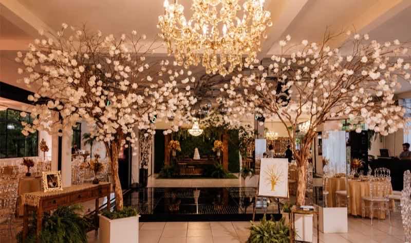 Salão iluminado com árvores de flores brancas, grande lustre dourado e mesas postas