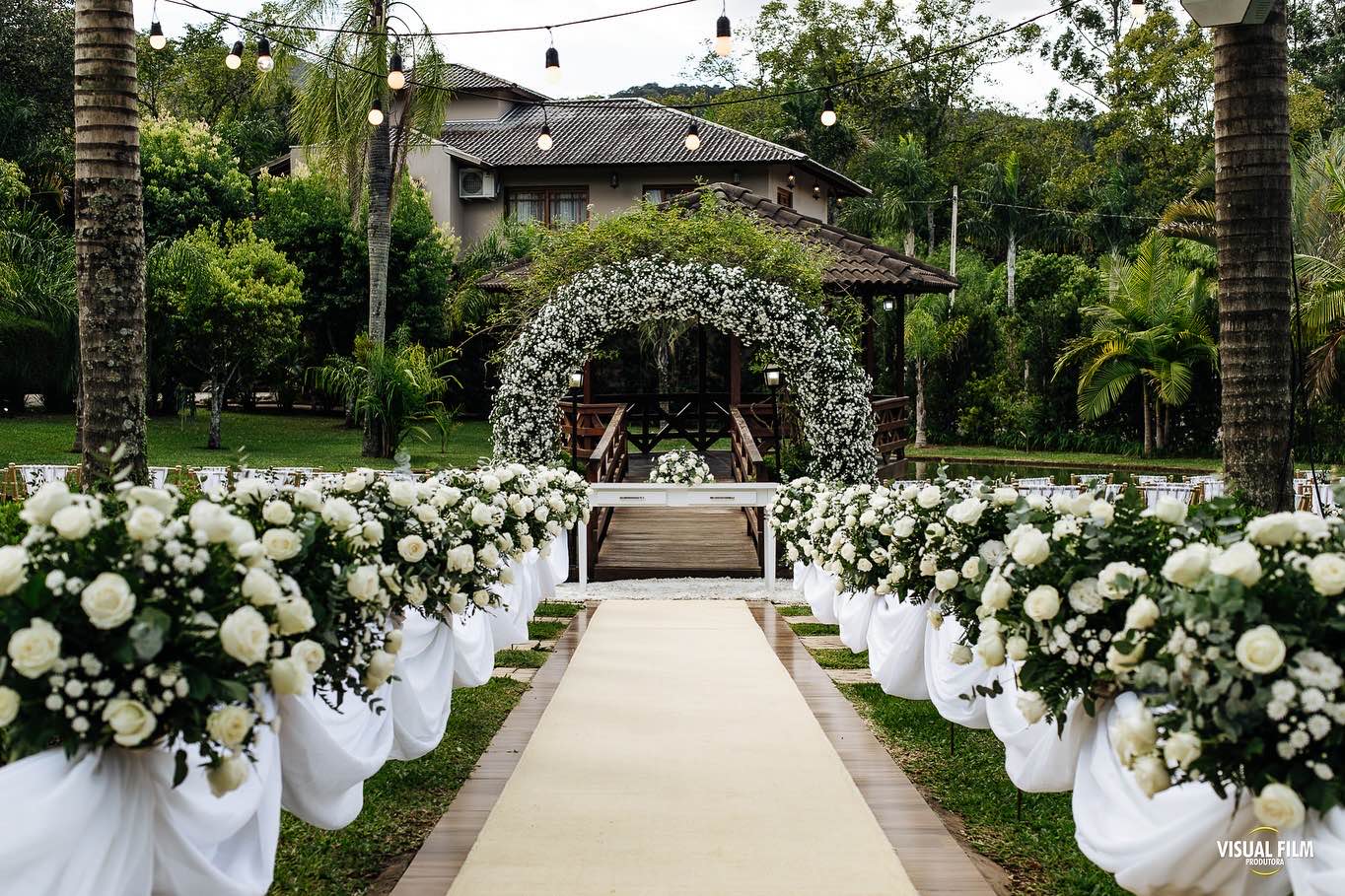 Corredor da cerimônia ao ar livre com arranjos de rosas brancas levando a um arco de flores diante do gazebo, entre palmeiras e varais de luzes